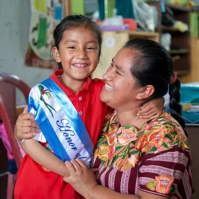 Niña sonriente con banda escolar abraza a su madre