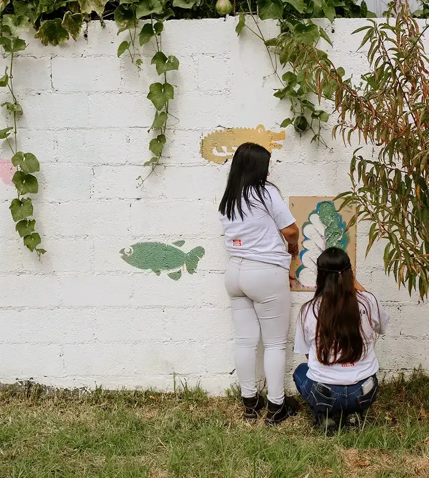Dos mujeres de espaldas pintan un muro blanco con peces y plantas trepadoras