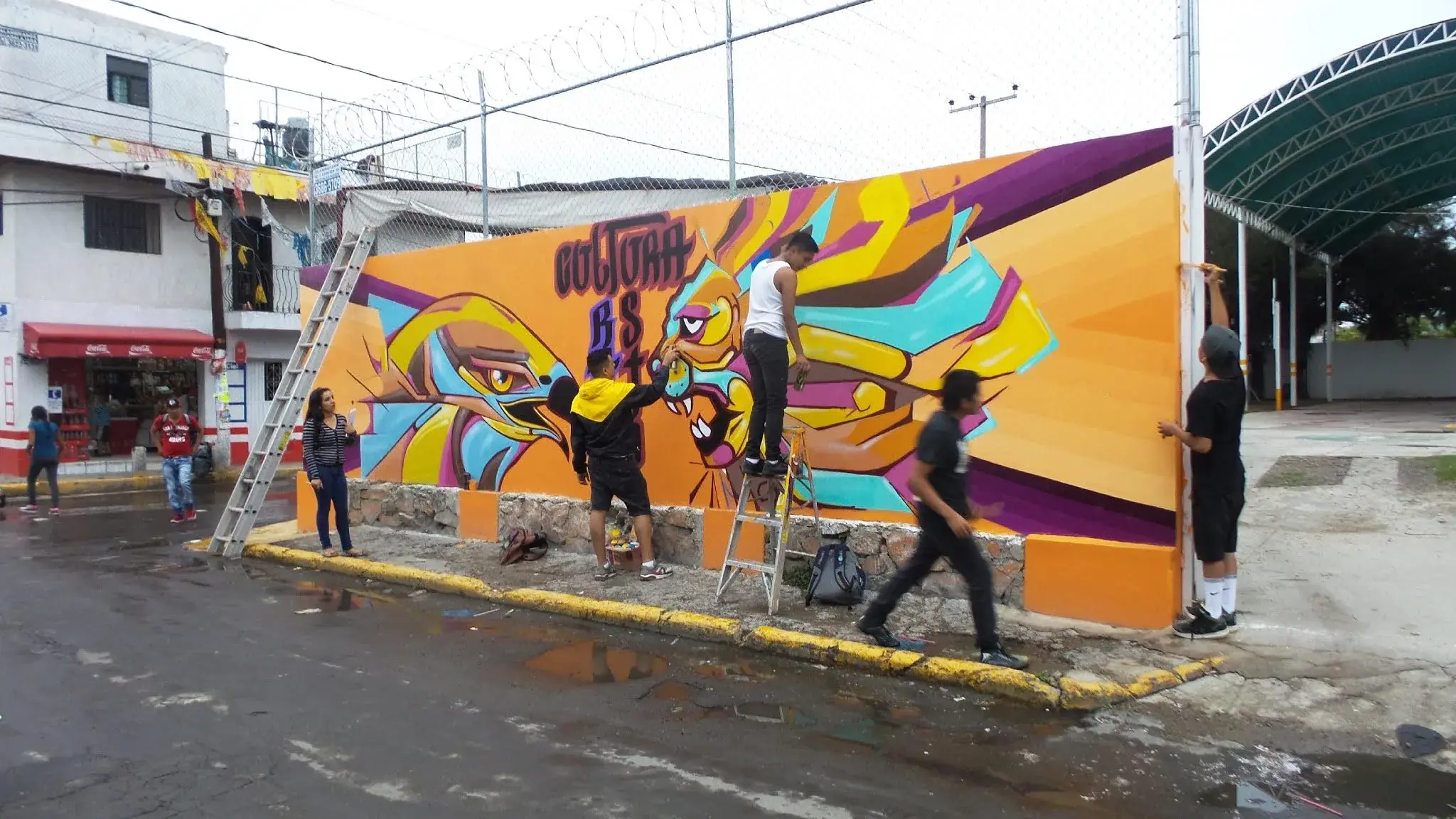 Jóvenes pintan un mural naranja y morado sobre una barda, con escaleras y andamios