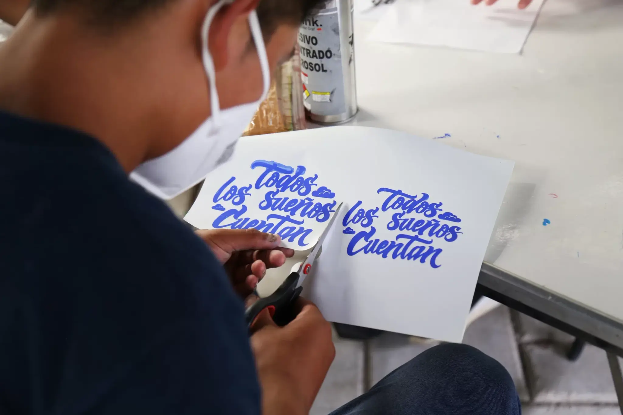 Un participante con cubrebocas recorta una hoja con el lema Todos los sueños cuentan en lettering azul