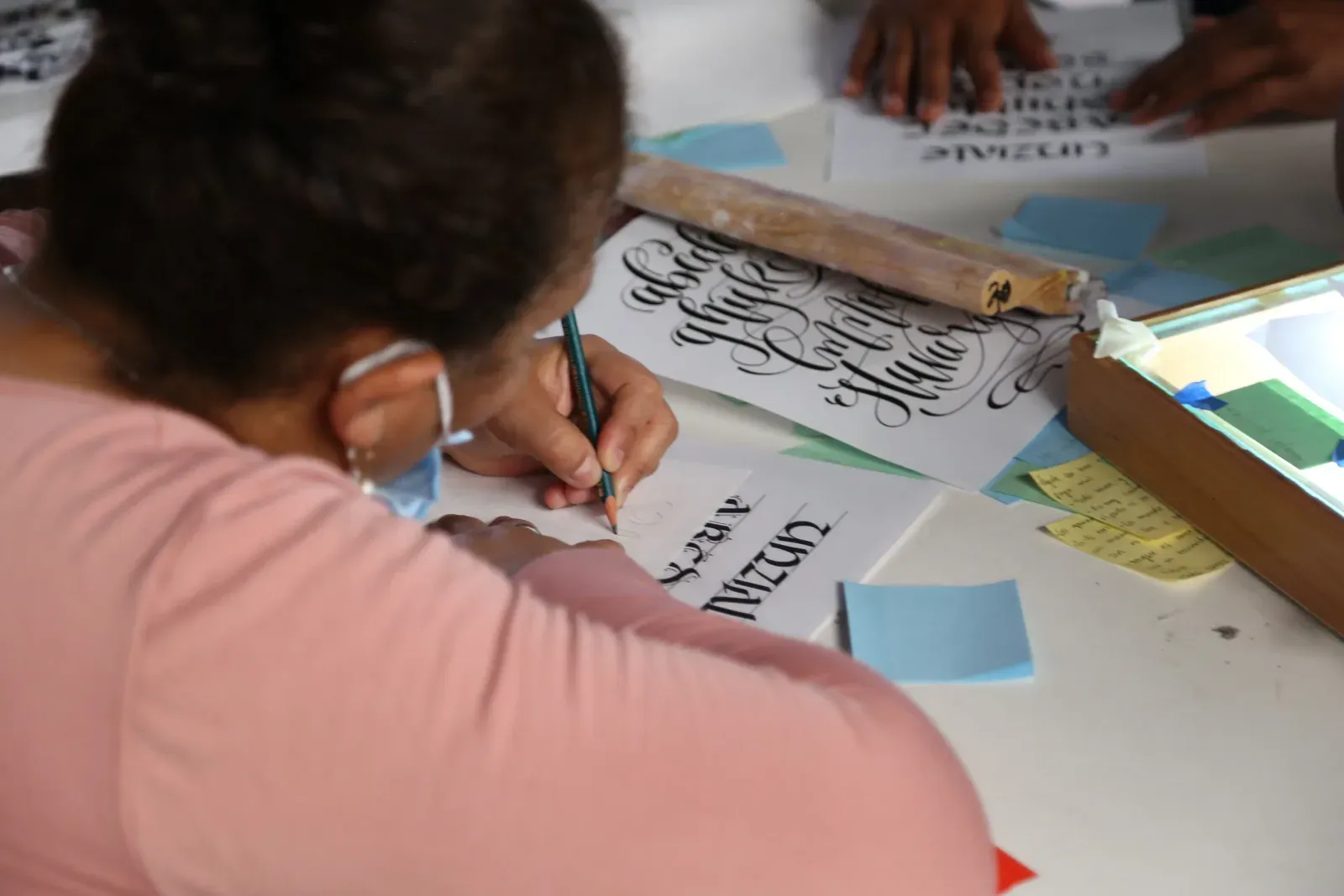 Una participante con cubrebocas escribe letras caligráficas sobre papel en una mesa de trabajo